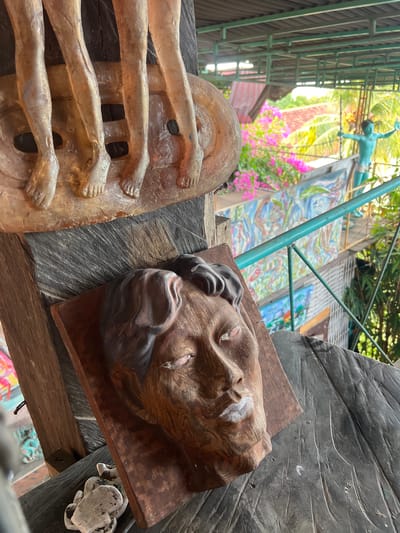 The image is a close-up of a collection of weathered wooden sculptures displayed outdoors in Tejakula, Indonesia. In the foreground, a carved wooden head with a stern expression and applied white paint on the lips is positioned on a dark, textured wooden surface. To the left and slightly above, a portion of a sculpture depicting several pairs of legs and feet emerges from a circular base. In the background, out-of-focus, there is lush greenery, pink bougainvillea flowers, and a turquoise metal railing. Further back, another turquoise sculpture of a human figure with outstretched arms is visible, along with colorful murals on a wall. The scene suggests an outdoor art installation or display, likely in a garden or courtyard setting, given the tropical foliage and open-air structure.