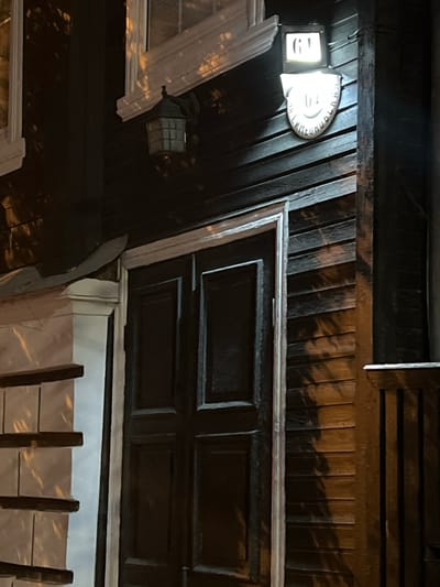 The image shows the exterior of a dark wooden building at night. Above a dark, paneled double door, there is a sign with the number "61" illuminated by a light fixture. Below this, a curved plaque with some text is visible, though it is difficult to read. To the left of the door, there is a window with white trim and a decorative molding. To the right of the door, a wooden railing is visible. The building appears to be a traditional structure, possibly a house or a historical building. The lighting suggests it is nighttime, and shadows are cast on the wooden facade. Without additional information or context, the specific location in Tver, Russia, cannot be determined from the image alone.