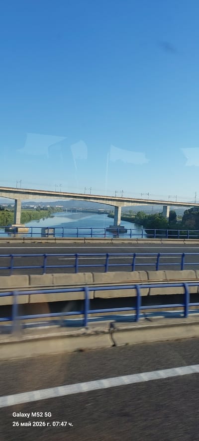 The image depicts a view from a bridge in l'Aldea, Spain. The photo was taken from a vehicle on the bridge. Another bridge spans a wide river with green vegetation on both banks. The sky is clear and blue. Safety railings line the bridge. The image has a timestamp indicating it was taken on May 26, 2026, at 07:47. The device used to capture the photo is a Galaxy M52 5G.
