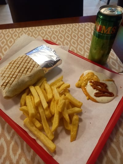 A red tray holds a wrapped sandwich, a pile of french fries, and a dollop of sauce. The sandwich is wrapped in foil. The fries are golden yellow. The sauce appears to be a mixture of white and orange with a drizzle of a darker sauce. A green can sits to the left of the tray. The tray is lined with white paper. The tray and its contents sit on a table with a patterned tablecloth.