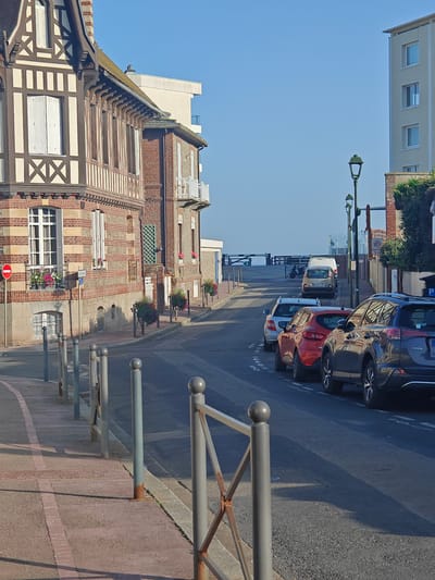 A view down a street in Étretat, France, leads towards the ocean. The street is lined with buildings, one of which is a half-timbered house with a red-tiled roof and white shutters. Another building is a modern, light-colored apartment building. Cars are parked along the right side of the street. The sky is clear and blue, suggesting a sunny day. The time of day appears to be morning or late afternoon due to the long shadows cast by the buildings and street furniture. The presence of a pedestrian walkway with bollards and a railing suggests a public area, possibly a promenade or street leading to a coastal viewpoint. There is a red and white circular sign with a horizontal bar on the left side of the street, indicating a prohibition. A sign with a green arrow and "P" is visible on the right. The scene evokes a calm, peaceful atmosphere.