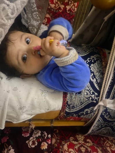 A baby lies upside down on a patterned surface. The baby is wearing a blue outfit with light gray trim around the cuffs and has dark hair and eyes. The baby is holding a yellow and purple object in its hands and has it near its mouth. The surface beneath the baby is patterned with white fabric that has light gray images on it. Behind the baby, there is a surface with blue and silver patterns on it. To the right of the baby, there is a surface with red and white floral patterns. The scene is indoors and well-lit. The location is Galaosiyo, Uzbekistan.