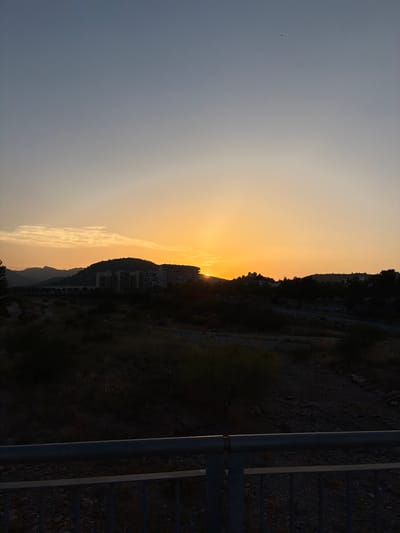 The image captures a sunset over the city of Sagunto, Spain. The sun is partially obscured by a distant mountain range, casting a warm orange glow across the sky. In the middle ground, several buildings, possibly apartments, are silhouetted against the vibrant sunset. The foreground is dominated by a dark, rough terrain, possibly a dried-up riverbed or a rocky area, with sparse vegetation. A dark metal railing runs across the bottom of the frame, suggesting the viewpoint is from a bridge or an elevated walkway. The sky transitions from a pale blue at the top to a bright orange near the horizon. There are no people visible in the scene.