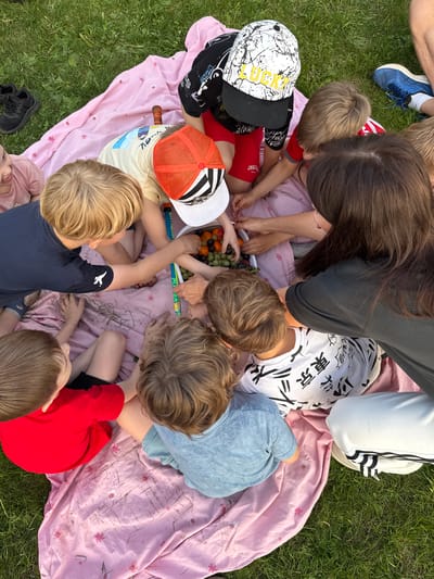 The image shows a group of children and one adult gathered around a pink patterned blanket on a grassy area. The adult, with long brown hair, is leaning over a bowl of fruit, possibly grapes and oranges, on the blanket. The children, all with fair hair, are reaching for the fruit. They are wearing various clothes, including t-shirts and shorts in colors like red, blue, and white with Japanese writing. Some are wearing hats. There are shoes visible in the periphery on the grass. The scene is likely a picnic or gathering. There is no indication of location in Кузнецово, Russia.