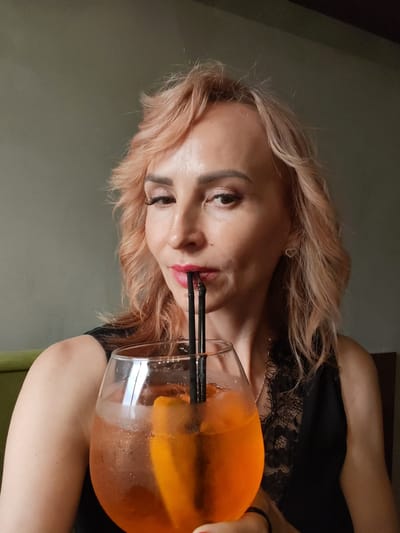 A woman with shoulder-length blonde hair and makeup is captured in a medium close-up, holding a large glass of an orange beverage with ice and an orange slice. She is using two black straws to drink from the glass. She is wearing a black top with lace detailing at the neckline and a necklace. The background is a plain, muted gray wall. No information is available regarding the city of Selivanovo, Russia in this image.