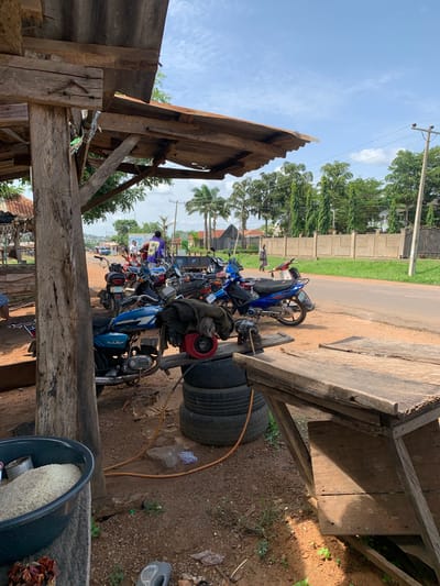 The photograph depicts a scene at a roadside stall in Gbeleko, Nigeria, on a bright, partly cloudy day. In the foreground, a large, rough-hewn wooden post supports a corrugated metal roof structure. Beneath the shelter, several motorcycles are parked on the dirt ground. A blue motorcycle with a black seat is prominently featured, with a worn black jacket draped over its rear. To the left, a dark basin filled with white rice sits on a wooden surface, next to some dried red objects. Stacked tires are visible behind the basin.
Beyond the immediate stall area, a paved road runs horizontally across the image. A few individuals are present on the road and sidewalk. One person in a bright purple jacket stands near a truck on the right side of the road, while another figure in gray walks further down the road on the left. A long concrete wall with a decorative iron gate runs along the right side of the road, bordered by green grass and tall trees, including palm trees. Power lines are visible against the sky. The overall impression is one of a busy, informal commercial area situated alongside a main thoroughfare in a Nigerian town.