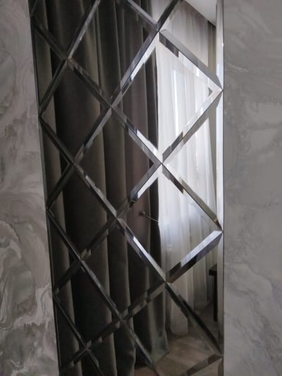 The image shows a wall adorned with beveled mirrors arranged in a diamond pattern, reflecting dark gray curtains and sheer white curtains in the background, suggesting an interior space. To the left and right of the mirrored wall are sections of marble-patterned tile. The scene is indoors and appears to be a residential or commercial setting. There are no people visible in the image. The location provided, Bezlesnyy, Russia, cannot be verified from the visual information presented in the image.