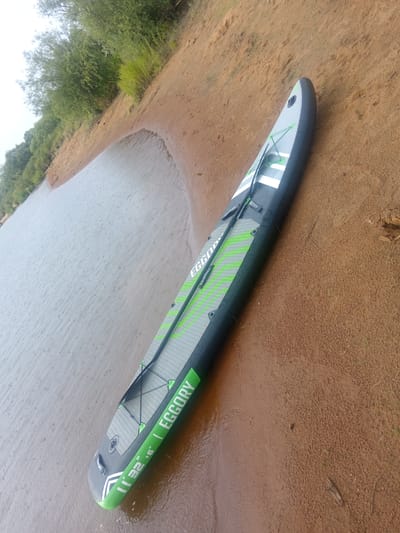 A dark gray and green stand-up paddleboard (SUP) lies on a sandy shore next to a body of water. The paddleboard is positioned diagonally, with its tail pointing towards the upper right corner of the frame. It is primarily dark gray with prominent lime green accents, including stripes along the sides and the brand name "EGGORY" printed in white on the green sections. The deck of the SUP features a textured gray surface with black bungee cords crisscrossing the front for cargo storage. The water appears calm with visible raindrops creating ripples on its surface. The sandy shore transitions into a vegetated embankment with green trees and grass in the background. The overall environment suggests an outdoor recreational setting, likely near a lake or river, possibly in Vasil'evka, Russia. There are no people or animals visible in the image.