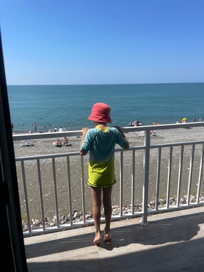 A young person stands on a balcony, facing a beach and the sea. They are wearing a red bucket hat and a two-toned t-shirt, with the top half turquoise and the bottom half lime green, and dark shorts. The beach is composed of pebbles and sand, with several lounge chairs scattered around. In the water, numerous people are swimming and wading, and some are on watercraft. The sky is clear and blue. The balcony has a white railing, and the floor is tiled. The overall setting suggests a sunny day at a resort in Giachrypsh, Abkhazia.