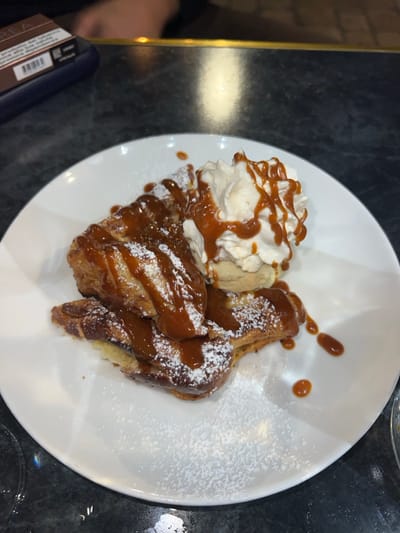 The image depicts a plated dessert served on a dark, mottled countertop, indicative of a restaurant or cafe setting in Orléans, France. The primary subject is a white, round plate presenting several pieces of golden-brown, layered pastry, possibly a type of French toast or a baked good, generously drizzled with a glossy, amber-colored caramel sauce. A light dusting of white powdered sugar covers both the pastry and portions of the plate. Adjacent to the pastry is a large dollop of white whipped cream, also topped with caramel sauce, resting on a small, round, light-colored biscuit or cookie. In the blurred background, to the upper left, a dark blue rectangular object with a brown label (possibly a tobacco product or similar item) is visible near a polished metallic strip, likely a bar rail or table edge. A blurred dark shape, consistent with a person's hand or arm, is partially visible behind these items. No specific human actions or interactions are visible beyond the presentation of the food.