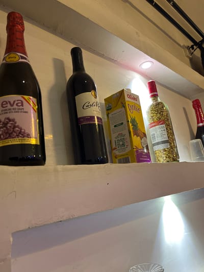 This image depicts a shelf displaying various beverage bottles and a juice box. The scene is indoors, likely a pantry or a display area in a store, given the shelf structure and lighting. The prominent objects include three bottles of wine, one juice box labeled "Chivita Qyotic" with pineapple and coconut imagery, and another bottle containing green olives. The bottles are arranged vertically on the shelf.
The bottle at the bottom is labeled "eva SPARKLING RED GRAPE" and appears to be a wine or sparkling beverage. Above it is a dark bottle of "Carlo Rossi SWEET RED WINE." The bottle containing green olives is a clear glass bottle with a red cap and a label indicating "STREET." The "Chivita Qyotic" juice box is positioned between the olive bottle and a red-capped bottle at the very top of the shelf.
Visible text includes brand names like "eva," "Carlo Rossi," and "Chivita," as well as product descriptions such as "SPARKLING RED GRAPE," "SWEET RED WINE," and "PINEAPPLE NECTAR." There are also indications of origin, such as "PRODUCT OF SPAIN." The lighting suggests it might be daytime or artificial light is being used in a dimly lit area. The overall impression is one of a stocked beverage shelf.