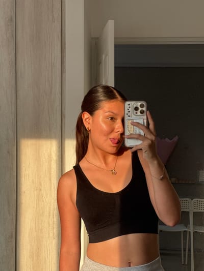 A woman is taking a selfie with a phone. She is wearing a black sports bra and grey sweatpants. The photo appears to be taken indoors, next to a wooden surface with strong sunlight casting shadows. A partial view of a window or a door is seen in the background. The scene doesn't provide enough context to locate it in Ocotlán.