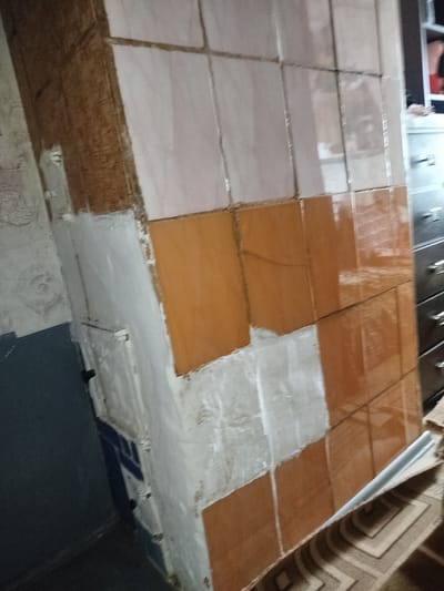 The image shows a partially tiled wall in what appears to be a residential or utility area in Уваравіцкі сельскі Савет, Belarus. The wall is constructed with a base of rough, possibly wooden material, which is then covered with various tiles. The upper section of the wall features light-colored, square tiles, possibly ceramic, with visible grout lines. Below this, a section of the wall is covered in warm-toned, square tiles, possibly also ceramic, with a glossy finish. There are areas of exposed concrete or plaster between the tiled sections and along the lower part of the wall, indicating ongoing construction or renovation. To the left of the tiled area, a dark blue surface is visible, possibly a painted wall or a piece of furniture. Further to the left, a textured wallpaper with a faint pattern can be seen. To the right of the tiled wall, a tall, dark wooden dresser or cabinet with multiple drawers is present, with metal handles visible. On top of the cabinet, a portion of a shelf is visible, containing some red items. The floor is covered with a patterned rug. The lighting suggests an indoor environment, possibly with artificial light. The scene depicts a work in progress in a domestic setting.