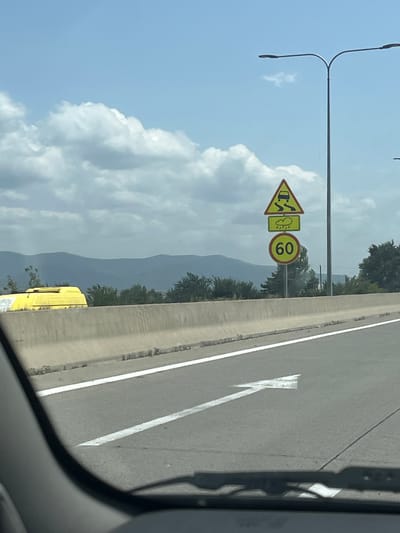 The image captures a view from a vehicle on a highway. In the foreground, the road surface is visible with a white lane marker and a directional arrow pointing left. A concrete barrier runs along the right side of the road. Further ahead, a set of traffic signs is mounted on a pole. The signs include a warning for slippery road conditions due to rain, indicated by a car skidding on a wet road and a cloud with raindrops. Below this, a circular sign displays "60" in red on a yellow background, signifying a speed limit of 60. In the background, distant mountains are visible under a partly cloudy sky. A yellow vehicle is partially seen behind the concrete barrier on the left side of the image. The scene appears to be an outdoor setting, possibly a highway environment, with no people clearly visible. The location, Kvemo Shavshvebi, Georgia, is not explicitly identifiable from the visual elements in the image itself, but the traffic signs suggest a regulatory context for road travel.