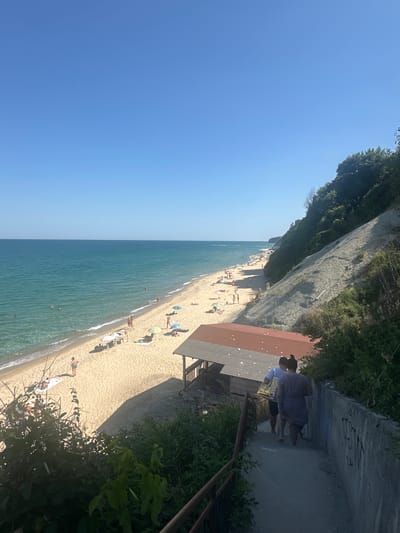 This is a photograph taken from a high vantage point looking down at a beach in Byala, Bulgaria. The scene depicts a sunny day at the Black Sea coast. The turquoise water meets a sandy beach dotted with people sunbathing and swimming. Umbrellas provide shade for some beachgoers.

In the foreground, a concrete pathway descends, with a railing on the left side. Two individuals are walking down the path, facing away from the camera. One is wearing a blue and white floral dress, and the other a white t-shirt and dark shorts. Behind them, a building with a reddish-brown roof is partially visible. To the right of the pathway, a steep, vegetated cliff face rises, covered in lush green trees and shrubs. Graffiti is visible on the concrete wall to the right of the path.

The sky is a clear, bright blue, indicating good weather. The overall impression is one of a popular summer destination, with people enjoying recreational activities by the sea.