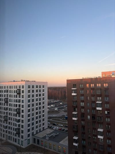 An elevated view captures a cityscape in Yanino-1, Russia, during golden hour, characterized by a clear sky transitioning from pale blue overhead to a warm orange-pink at the horizon. The scene is dominated by two modern, multi-story residential buildings.

To the left, a tall, light grey or white brick building rises, featuring numerous uniformly-sized, rectangular dark-framed windows arranged in a grid pattern. Several windows incorporate dark, projecting, enclosed balconies. A lower, flat-roofed section with large, reflective windows, some displaying red markings, extends from its base in the foreground.

To the right, a multi-story building constructed of dark red or brown brick is partially visible. This structure also features numerous rectangular windows, many equipped with small white projecting balconies or window boxes. The building's windows reflect the warm hues of the horizon.

Between these two buildings, an asphalt parking lot is visible, accommodating numerous parked cars of various colors, predominantly dark and white. Beyond the parking area, a construction site with scattered white vans and heavy equipment is discernible. The background features a dense, bare deciduous forest stretching across the horizon, illuminated by the low sun. The immediate foreground shows a paved, dark grey surface with a curved edge. No individuals are visible within the frame.
