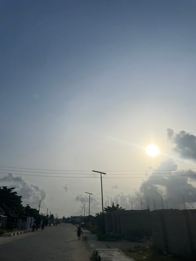 The image depicts a street scene in Mosafejaw, Nigeria, under a partly cloudy sky with the sun prominently visible in the upper right quadrant. The sky transitions from a light blue-grey at the top to a hazy yellow-white near the bright, circular sun, which emits a distinct lens flare. Scattered cumulus clouds, ranging from white to dark grey, are present across the mid-ground of the sky.
A paved road, constructed from interlocked pavers, extends from the foreground into the distance, exhibiting a slight curve. Overhead power lines span horizontally across the mid-ground. Along the left side of the road, a series of single-story structures are partially visible behind green foliage.
On the right side of the paved road, a dirt path runs parallel. Multiple dark-colored streetlights, each equipped with an integrated solar panel, stand vertically along this path, receding into the background. Adjacent to the closest streetlights, a partially constructed building with exposed rebar is visible.
Several individuals are present on the road, silhouetted against the bright lighting. One person, dressed in dark attire, walks away from the viewer on the right side of the paved road. Additional human figures are discernible walking further in the distance. The scene is characterized by strong backlighting from the low sun, creating significant contrast.