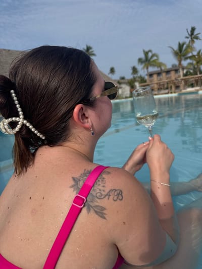 A single female individual is depicted from the back and right side, seated within a light blue swimming pool under a clear sky. Her dark brown hair is pulled back into a low bun, secured by a large white pearl claw clip. She wears round, gold-rimmed sunglasses and a blue drop earring on her right ear. A bright pink swimsuit strap is visible on her left shoulder, adorned with a pink adjustment buckle. A large, dark-ink floral tattoo occupies her upper left back, with the initials "A B" tattooed below it. Her skin has visible moles and appears wet from the pool. Her right hand holds a clear stemmed glass containing a light-colored liquid, likely a beverage. A delicate silver bracelet is visible on her right wrist. In the background, beyond the pool, are multiple palm trees and low-rise buildings with thatched roofs, characteristic of a resort in Zanzibar, Tanzania. The sky is bright blue with scattered white clouds.