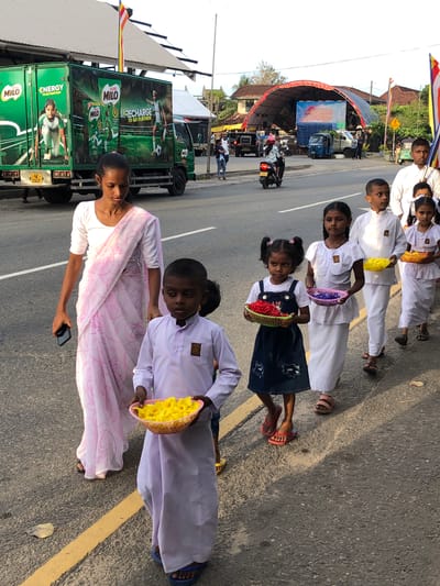 The image depicts a street scene in Habaraduwa, Sri Lanka, likely during the daytime given the bright lighting. A group of people, including children, are the primary subjects. The children are dressed in white robes or uniforms and some are holding baskets filled with what appear to be yellow flowers. One child, wearing a blue dress over a white top, also holds a colorful basket. A woman in a pink and white patterned sari stands in the foreground, looking down at her phone.
Behind the people, a large green truck with "MILO" branding is visible, suggesting a promotional event or parade. The truck features images of athletes and the slogan "ENERGY TO GO FURTHER." Another truck with a large curved red awning is also present, and a small three-wheeled auto-rickshaw is parked nearby. Buildings with traditional architecture are visible in the background. The ground is asphalt, marked with yellow lines.
The overall atmosphere appears to be festive and possibly part of a cultural or religious procession given the attire of the children and the presence of a Buddhist flag flying from the Milo truck. The children's expressions are neutral, while the woman in the foreground seems focused on her phone. The weather appears clear.