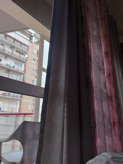 The media file presents an interior view looking outwards through a window, captured at a slightly upward angle. In the upper portion, partially drawn roller blinds or shades are visible, featuring a distinct floral pattern in shades of pink and white with dark outlines. Below these blinds, a dark, horizontal beam or ledge, possibly made of wood or metal, spans across the middle foreground, partially obstructing the view outside.
Through the window's multiple panes, an urban landscape unfolds. Several multi-story residential buildings, primarily in tones of beige or light orange, are visible. These structures feature numerous windows, balconies, and some external air conditioning units. On the far right, a dark, intricately carved or cut-out decorative element, possibly a piece of furniture or part of an interior architectural feature, is partially visible.
The scene takes place during the daytime, evidenced by the bright natural light illuminating the exterior buildings and the clear weather conditions. No specific activity or event is occurring, and the image captures a static moment. There is no discernible visible text. The location context for this urban scene is Tiranë, Albania.