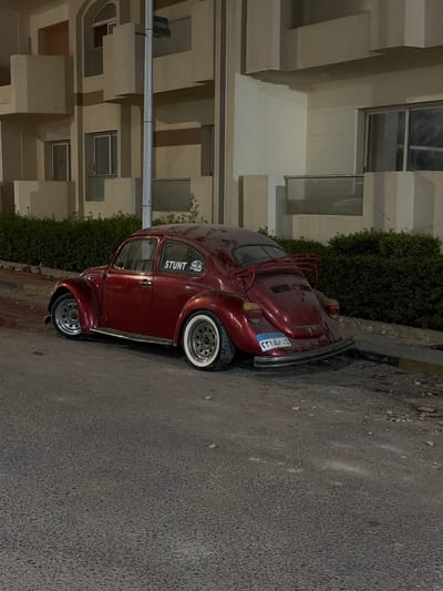 A red Volkswagen Beetle is parked on a paved road in Hurghada, Egypt. The car has a luggage rack on the back. A sticker on the driver's side door reads "STUNT" with a logo. The car is parked in front of a building with multiple stories and balconies, and a hedge runs along the side of the road. The scene appears to be at night, as indicated by the lighting conditions.