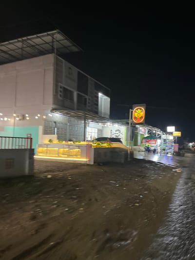 The image depicts a street scene at night in Landuh, Indonesia, possibly after rain, as indicated by the wet, reflective surface of the ground. The scene is illuminated by various lights, including those from buildings, signs, and string lights, creating a vibrant atmosphere.
The main subjects are buildings with modern architecture, featuring white exteriors, balconies, and metal scaffolding. One building appears to be a multi-story establishment with an open-air dining or seating area on the upper floors, illuminated by warm, yellow string lights. Another building, visible on the left, has a more industrial look with a large metal structure and dark facade.
There are several signs visible. A prominent sign in the center features a circular logo with a yellow design and orange neon outlines, possibly representing a restaurant or establishment. Another sign above it is a rectangular white board with illuminated text, but it is difficult to read clearly. Further down the street, more illuminated signs with colorful lights suggest a commercial area.
Although the image is blurry, it's possible to discern a few figures that appear to be people in the background, sitting at tables or walking. The overall activity seems to be commercial, with the buildings likely housing shops, restaurants, or other businesses that are open at night.
Notable details include the time of day being night, the wet ground suggesting recent rain or a damp environment, and the bright, artificial lighting that contrasts with the dark sky. The location context of Landuh, Indonesia, provides a geographical anchor for the scene. The atmosphere feels lively and bustling, despite the darkness.