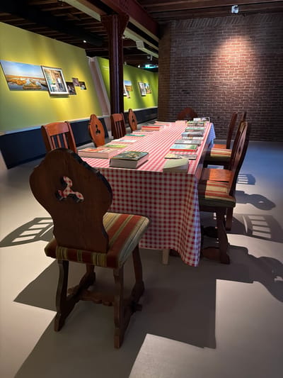 The image shows a long table covered with a red and white checkered tablecloth, set in a room with exposed brick walls and wooden beams. The table is laden with books, some stacked and others open. Around the table are several wooden chairs with upholstered seats. The room appears to be an exhibition space, with framed photographs displayed on a lime green wall in the background. One photograph depicts a scenic landscape with water and reeds, and another shows a person standing in an outdoor setting. The lighting in the room is dramatic, casting long shadows from the chairs onto the floor. This scene is located within the Maritime Museum in Rotterdam, Netherlands, which houses a diverse collection of artifacts and exhibits related to seafaring history and maritime culture.