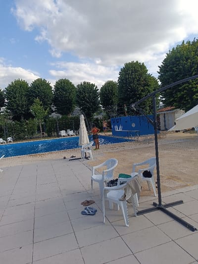 A sunlit outdoor swimming pool area features a bright blue pool surrounded by a tiled terrace. White plastic chairs are scattered around, some with personal items like towels and sandals. People are visible near the pool, including a person on a diving platform. Trees line the area beyond the pool, and a cloudy sky is overhead. A large umbrella stand is prominently positioned in the foreground.