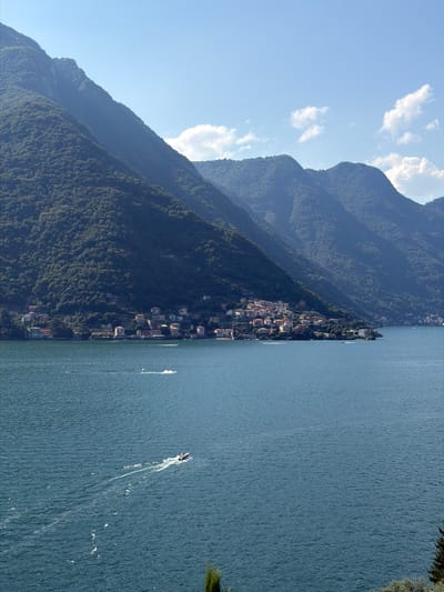 The image depicts a wide view of Lake Como, with the town of Pognana Lario situated along its shoreline, nestled at the base of steep, tree-covered mountains. The water is a deep blue, with a few small boats creating white wakes. The town itself is a cluster of buildings with red-tiled roofs, seemingly clinging to the mountainside. The sky is a bright blue, with a few scattered white clouds. The overall impression is one of a serene, picturesque landscape.