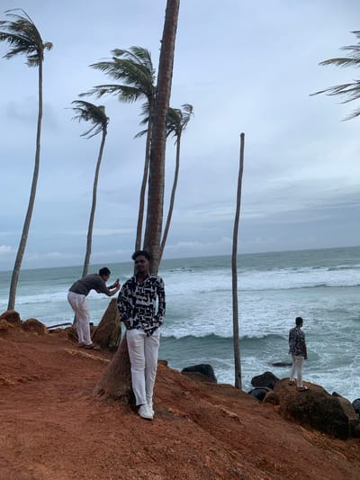 The image depicts a group of people near the ocean in Thalaramba, Sri Lanka, on a cloudy day. The primary subject is a man in the foreground, dressed in a black and white patterned shirt and white pants, standing on a reddish-brown rocky cliff overlooking the sea. He is leaning against a palm tree, looking directly at the camera with a neutral expression.
Behind him, another person is visible, also wearing white pants and a grey t-shirt, holding up a phone as if taking a picture. Further in the distance, a third person in a similar outfit is seen standing on the rocky outcrop. The scene is characterized by several palm trees with their fronds swaying in what appears to be a breezy atmosphere. The ocean is turbulent, with waves crashing against the shore. The sky is overcast, suggesting a potentially humid or rainy climate common in Sri Lanka. The overall mood is casual and relaxed, with the individuals enjoying the coastal scenery. There is no visible text in the image.