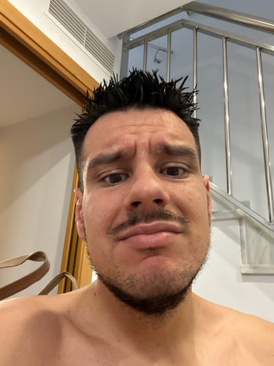 The image is a selfie taken indoors. In the foreground, a man with dark, spiky hair and a mustache is looking at the camera with a worried expression. His eyebrows are furrowed, and his mouth is slightly open. He is shirtless, and his shoulders and collarbone are visible. Behind him, to the left, a wooden door frame is visible with a white air conditioning vent above it. To the right, a modern staircase with metal railings and white steps can be seen. The background appears to be a residential interior, possibly a hotel or a home, in Roquetas de Mar, Spain. The lighting is bright, suggesting it's daytime. There are no other people or significant objects in the frame besides the man and the interior elements.