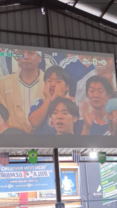The image shows a large screen displaying a soccer match with the score 0-0 between Japan and Sweden. On the screen, several people are visible watching the match. The people appear to be a mix of adults and children, all appearing to be of East Asian descent. The screen is mounted outdoors, under a roof. In the foreground, below the screen, there are several banners and posters. One banner is in Indonesian and appears to be advertising a soccer event. Another poster displays a man in a suit. The overall scene suggests a public viewing of a soccer match in an outdoor setting. The city of Bireuen, Indonesia is mentioned in the prompt, suggesting this is the location.