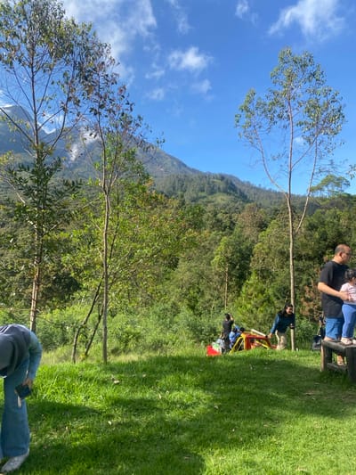 The image shows a verdant landscape with a group of people gathered on a grassy knoll, overlooking a lush, forested hillside. The hillside is characterized by a mix of evergreen trees, with a distinct line of taller trees forming the peak of the mountain. The sky above is a clear, bright blue with a few scattered white clouds.

A red and yellow vehicle is parked on the hillside, partially obscured by trees and vegetation. Three people are sitting in the vehicle, while a fourth person stands beside it, possibly engaging in conversation. 

On the grassy knoll, several individuals are present. One person, wearing a blue hooded jacket, is bending down, seemingly examining something on the ground. To the right of the vehicle, a woman is standing near the vehicle, her back to the viewer, while another person is seated in the grass. 

To the far right of the image, a man wearing a dark blue shirt and jeans stands beside a small wooden table. He is speaking with a young child wearing a pink shirt.

The image is captured in a rural environment in Kecamatan Tawangmangu, Indonesia, located at coordinates -7.661 latitude and 111.184 longitude. The scene suggests a leisurely outing, with the group enjoying the scenic mountain vistas and perhaps engaging in conversations or activities.
