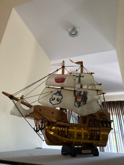 This media file depicts a model of the Santa Maria ship, likely a display piece. The model is predominantly made of wood, with white fabric sails that feature various emblems and designs. One sail has a green cross, another displays a coat of arms with an eagle, and a third shows a blue crest with lions. The ship has intricate rigging made of dark string, and two small anchor ornaments are attached to its side. The hull of the ship is detailed with yellow banding. The model is shown mounted on a white wall, with a black bracket labeled "SANTA MARIA" visible on the right side. The background includes a window with blinds and what appears to be a light fixture on the white wall to the left. The scene is indoors and well-lit. The overall impression is that of a well-crafted decorative item.
