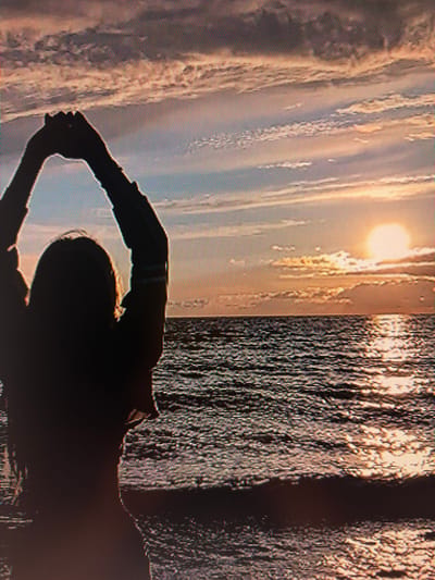 The image depicts a silhouette of a person with their arms raised above their head, standing on a beach at sunset. The sun is low on the horizon, casting a warm glow over the water and sky. The water is dark and rippling, with reflections of the sunset visible on its surface. The sky is filled with clouds, painted in hues of orange, pink, and blue.
Regarding the location in the city of Szczecin, Poland, the image provides no visual cues or identifiable landmarks to confirm this. The scene is generic and could be any coastal area during sunset. Therefore, it is not possible to determine the specific location within Szczecin based on the provided image.