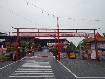 The image depicts the entrance to "Waroeng Sumringah Merdeka Bermain," located in Tanjungarum, Indonesia. A large, red archway with upward-curving eaves spans the entrance, displaying the establishment's name in white capital letters. String lights are suspended above the archway and extend into the interior area.

The foreground is paved with dark grey interlocking pavers, featuring white zebra crossing markings on the left approach and white dashed lines on the right. To the left of the main path, within the entrance, are low planters with black and white patterns containing red decorative elements, including two red parasols and a sign prohibiting food/drinks and pets.

On the right side of the entrance, a small building with a reddish-brown tiled roof and light-colored walls displays multiple information signs, some in Indonesian, including "LOKET MERDEKA BERMAIN." A yellow three-wheeled vehicle, resembling an auto-rickshaw, is parked facing away from the viewer, positioned just beyond the archway on the right.

Beyond the entrance, an open-air area is visible, furnished with multiple red tables, chairs, and parasols. Additional red decorative objects, including a red flamingo statue, are interspersed throughout. Several individuals are present within this area; one person in a blue garment is distinctly visible in the mid-ground. The background consists of green foliage and distant structures under an overcast, grey sky.