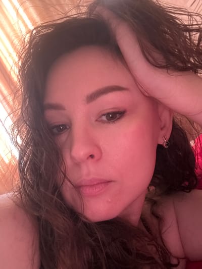An indoor close-up shot features a person, likely a woman, with dark brown, wavy hair, looking directly at the camera. Her head is tilted, giving the impression she is lying down. Her right eye (from her perspective) is clearly visible, with the left eye partially obscured by hair. She has neatly groomed eyebrows and appears to be wearing light makeup. Her lips are slightly parted.

An earring, possibly a small dark stud, is visible in her right ear. Her left arm is raised, with her hand resting near her forehead, partially covering the upper part of her head and hair. The skin tone has a warm, reddish-pink cast, which might be due to lighting conditions or a photographic filter.

The background is softly blurred, showing indistinct textures and colors. In the lower left corner and extending across some parts of the image, diagonal stripes of warm, yellowish-orange light are visible. These suggest sunlight filtering through horizontal blinds or a window, possibly indicating either early morning or late afternoon.

The person's expression is calm and relaxed, as she poses for the photograph. The setting appears to be a domestic space, such as a bedroom or living room. There is no visible text in the image.