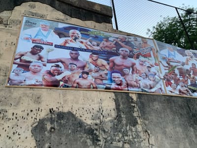 A large billboard is mounted on a concrete wall in Sokoto, Nigeria, at the coordinates 13.049 latitude, 5.238 longitude. The billboard is covered in various images of men in various poses, mostly in boxing-related clothing. The billboard is weathered and has a number of cracks and some missing pieces. A portion of the billboard features a collage of several images: In the upper left corner, an image of a man in a traditional white robe and a white cap, the top half of a white car, and a banner that says "DR. AHMAD ALIYU MUSAH." Below this are multiple images of young men wearing red boxing gloves and a small banner that says "DAMBEN MOTA NA SOKOTO." Below this, there are multiple images of young men wearing boxing gloves, some wearing headgear, others with bare heads. The bottom row features a small banner that says "MUSH" and a number of men and boys in boxing gear. The upper right corner features a banner that says "SOKOTO GIDA DA AHMAD TAMBUWAL" and multiple images of young men wearing boxing gloves and one man with a microphone. The middle right features a banner that says "BAHAGON ZAKI YANU IN ACTION" and features a man wearing boxing gloves and several other young men in the background. The lower right section features a banner that says "AHMAD ALIYU" and a number of images of young men wearing boxing gloves, most of whom are wearing red boxing gloves and shorts. The entire billboard is surrounded by a black metal fence. The background behind the billboard is a light blue sky with a few white clouds. There are no other people or objects in the image.