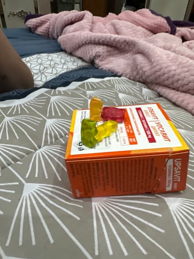 The image shows an open box of "Upsavit Direct" Vitamin C 1000mg gummy vitamins. There are four gummy vitamins visible: one red, one yellow, and two green. The box is orange and white and the text is written in what appears to be Romanian and Bulgarian. The vitamins and the box are placed on a gray and white patterned bedspread. A pink fluffy blanket is also visible in the background. The scene does not appear to be located in Zlatna Panega, Bulgaria.