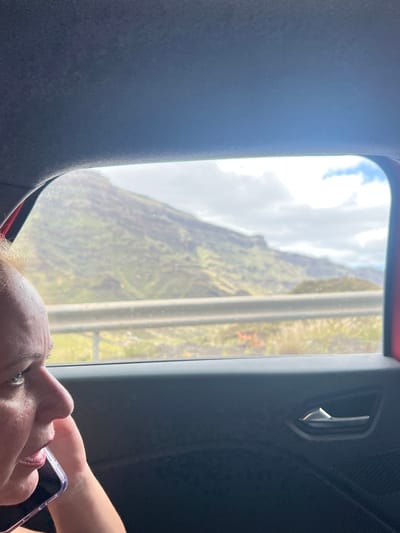 The image, taken from inside a vehicle, shows a person looking at a phone with a mountainous landscape visible through the window. The location is likely in Mogán, Spain, suggested by the arid, volcanic mountains characteristic of the Canary Islands. The window features a metal handle and a grey surround. The interior of the vehicle appears to be dark, and the person's face is visible in the lower portion of the frame.