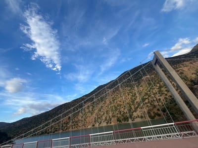 The image depicts a bridge in Ait Mazigh, Morocco, spanning a body of water. The bridge appears to be a suspension bridge, with a large concrete tower supporting the cables. A steep, arid mountain rises directly behind the bridge. The mountain is sparsely covered with vegetation. The sky is blue with scattered clouds. The railing of the bridge is a combination of red and white. The bridge deck is a dark brown.
