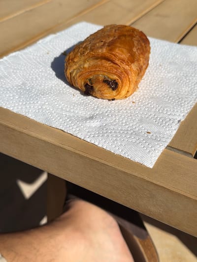A single, golden-brown pain au chocolat with a flaky, layered texture rests on a white, dimpled paper napkin. The napkin is positioned on a light-colored, horizontally slatted wooden surface, exhibiting visible grain patterns. Direct, bright sunlight illuminates the scene, casting a distinct shadow of the pastry onto the napkin and wood. Below the wooden surface, in the lower right quadrant, a portion of a light-skinned, hairy human foot wearing a dark sandal or shoe is partially visible. The scene is outdoors in Mougins, France.