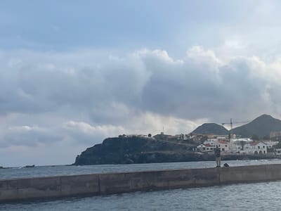 The image depicts a coastal scene in Cabo De Palos, Spain. In the foreground, a concrete pier or breakwater extends into dark, choppy water. A single individual, dressed in dark upper wear and light-colored trousers, stands on the pier holding a fishing rod, actively engaged in fishing. A backpack or bag is positioned on the pier near the individual.
In the mid-ground, a rugged, dark rocky headland juts into the sea from the left. Beyond the pier and headland, a coastal town is visible, characterized by white and light-colored buildings with red or orange tiled roofs, situated on rising terrain. Further in the background, dark, rounded hills are present, with a tall yellow construction crane standing prominently against one of the hills. The sky is overcast with numerous light grey and white clouds, though patches of lighter blue sky are visible in the upper sections.