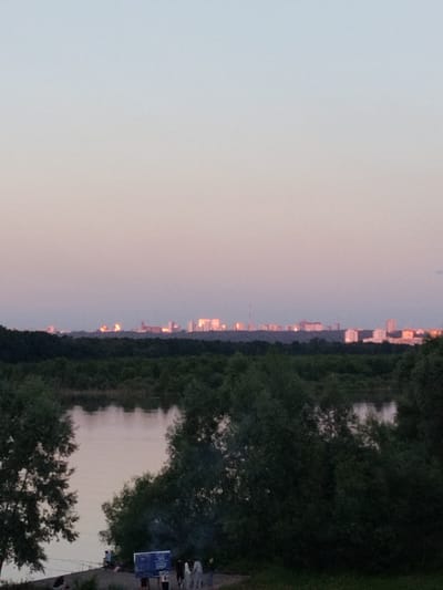 The image captures a distant cityscape with numerous buildings and structures visible on the horizon, bathed in the soft, hazy light of either dawn or dusk. A wide river or body of water occupies the foreground, with its surface reflecting the muted colors of the sky. Dense green foliage, consisting of trees and bushes, flanks the water and forms a dark, textured band across the middle ground, separating the water from the distant urban skyline. In the lower right portion of the frame, a group of people are gathered on what appears to be a riverbank or elevated area. One individual is seated, while others stand and appear to be interacting. A sign or board is also present in this area. The overall scene suggests a tranquil evening or morning view from a natural vantage point overlooking a city. The location is identified as Mikhailovka, Russia.
