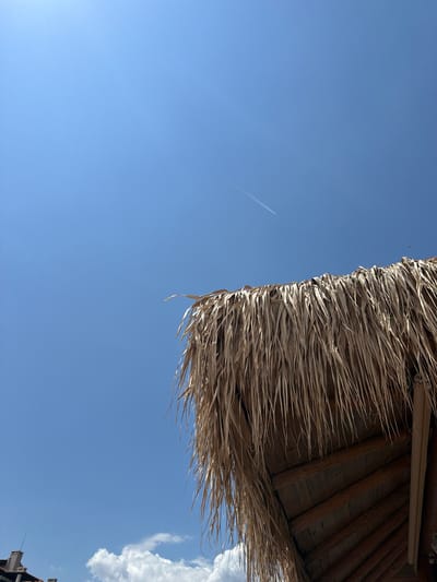 This is a low-angle shot of a thatched roof against a clear blue sky. A contrail from an airplane is visible in the upper right quadrant of the sky. The thatched roof is made of dried palm leaves and is attached to a wooden structure. In the lower left corner of the image, the rooftops of buildings with red tiles are visible, suggesting an urban or semi-urban environment. The overall impression is a sunny day, likely in a warm climate. This scene could be from the city of Lozenets, Bulgaria, known for its beaches and resorts.