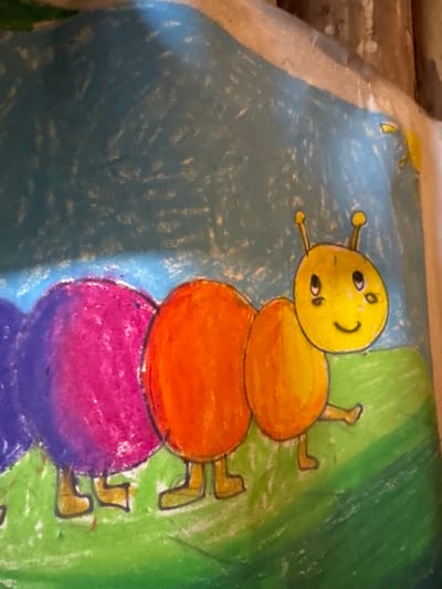 A child's drawing shows a multicolored caterpillar against a backdrop of blue and green. The caterpillar consists of a yellow head with a smiling face and two antennae, followed by a yellow, orange, pink, and purple rounded body segments. The caterpillar has small brown feet. The left side of the drawing is a light blue, while the right side is green. The drawing is likely on paper or canvas. It does not appear to be located in Mèo Vạc Commune, Vietnam, as the context is a child's artwork and there are no discernible elements to indicate such location.