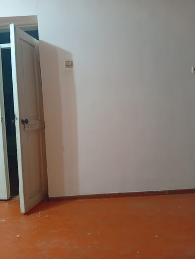 A wide-angle, slightly high-angle interior shot of an empty room shows a partially open white door on the left, revealing a glimpse of a darker interior. The door is adorned with a dark, round doorknob and decorative paneling. The interior wall is painted a light, neutral color, and the floor is a distinctive, bright orange. A dark brown baseboard runs along the bottom of the wall. In the upper left quadrant of the wall, a beige electrical outlet is visible. The scene is devoid of people and activity, suggesting an unoccupied space within a residence in the village of Zhartytobe, Kazakhstan.