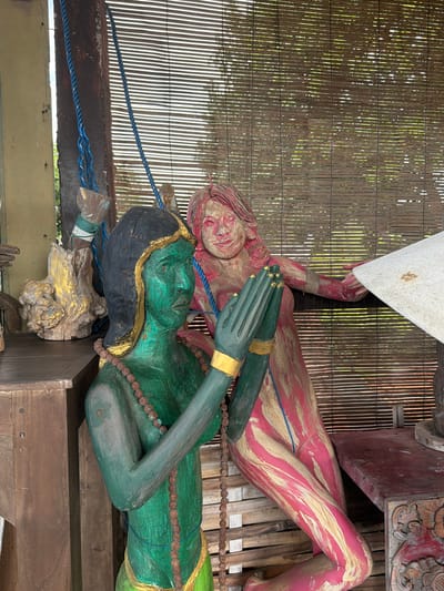 The image depicts a wooden sculpture of a woman painted green, with a gold headdress and necklaces, positioned on the left. Her hands are clasped in front of her chest, and she is adorned with a beaded necklace. To her right, another wooden sculpture of a woman, painted in pink and gold, leans to the right, with her left arm extended towards a lampshade. The background features a woven bamboo screen, and to the left, a wooden table and a piece of driftwood with gold accents. This scene is located in Tejakula, Indonesia.