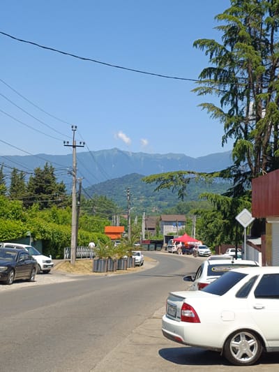 The image depicts a roadside scene in Vesyoloye, Russia, under a clear blue sky. In the foreground, a white sedan is parked on the right side of the asphalt road, with its rear facing the camera. Further down the road, other vehicles are visible, including a dark-colored sedan on the left, and a white SUV. A red canopy structure is set up on the left side of the road, with people gathered around it. Behind the road, there are buildings, lush green trees, and in the distance, rolling hills and mountains. Electrical poles and wires are strung across the scene, indicating infrastructure in the area. The overall impression is a somewhat rural or suburban environment on a sunny day.