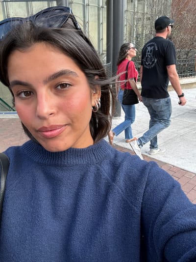 The image depicts a close-up selfie of a young adult female situated on a city sidewalk. She has dark brown hair, dark eyes, rosy cheeks, and is wearing a navy blue mock-neck knit sweater, silver hoop earrings, and sunglasses resting on her head. Her expression is neutral, and she is looking directly at the camera.
In the midground, two individuals are walking from right to left on a reddish-brown brick sidewalk. The male wears a black baseball cap, a black t-shirt with a white graphic on the back featuring "PROPRIETORS OF..." and "HERITAGE" text, faded blue jeans, and black-and-white checkered Vans slip-on shoes. He has a dark watch on his left wrist and holds a cylindrical object in his left hand. The female walks beside him, wearing a red t-shirt, blue jeans, tan flat shoes, and carries a black shoulder bag. She has curly hair and wears glasses.
The background features a large building with a distinctive glass and metal facade, characteristic of modern public architecture. Partially visible text in the upper left indicates "Regional Rail." This specific architectural style combined with the "Regional Rail" signage confirms the location as the vicinity of 30th Street Station in Philadelphia, Pennsylvania, United States. Bare trees are visible in the distant background.